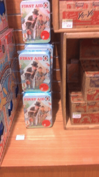 A display shelf featuring tins labeled "First Aid" with retro-style illustrations of children helping each other with basic first aid. Each tin has a red circle labeled "Activity Kit 0" and is stacked neatly. Adjacent are wooden boxes containing vintage-style product packaging, including yo-yo sets.