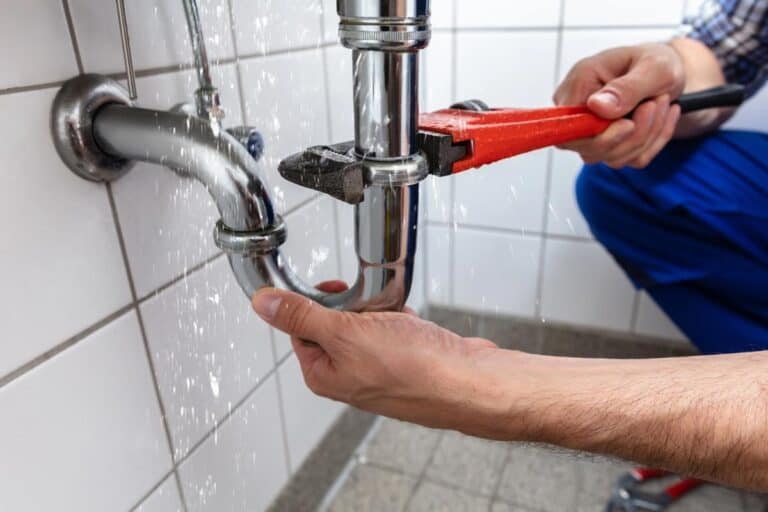 A professional plumber using a wrench to repair a leaking sink pipe, with water spraying from the joint. This highlights the benefits of hiring a licensed plumber to handle complex plumbing issues safely and efficiently, preventing costly water damage.