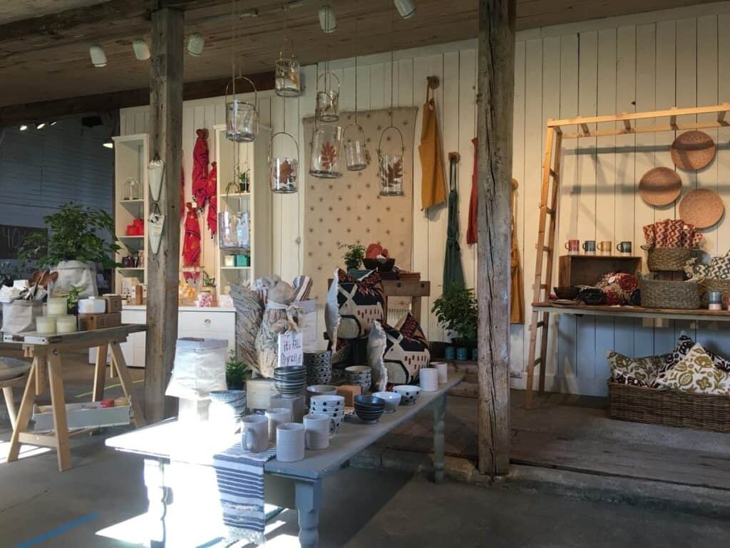 A rustic home decor store interior featuring a mix of bohemian and vintage-inspired pieces. The space includes wooden beams, white-paneled walls, and an assortment of textiles, woven baskets, ceramics, and glass lanterns. A cozy collection of throw pillows, aprons, and decorative items is arranged on tables and shelves, creating a warm and inviting aesthetic.