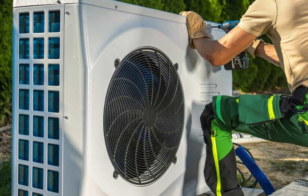 A technician wearing work gloves and green-and-black work pants is installing an outdoor energy-efficient heat pump unit. The unit features a large black fan and a protective grid on the side. The technician is using a power drill to secure components while connecting blue insulated pipes. The background includes green shrubs, indicating an outdoor residential or commercial setting. This image represents the installation of an efficient heating and cooling system designed to optimize home energy use and reduce costs.