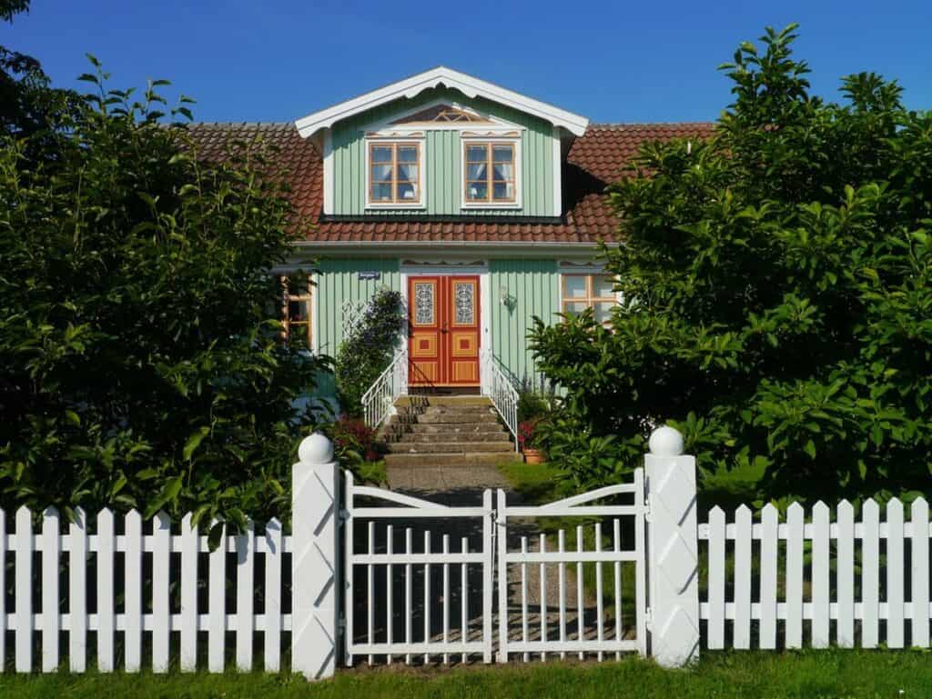 A light green house with red accents framed by a bright white picket fence and gate, with lush greenery on both sides. Classic picket fencing adds charm and curb appeal while creating a welcoming front yard boundary.