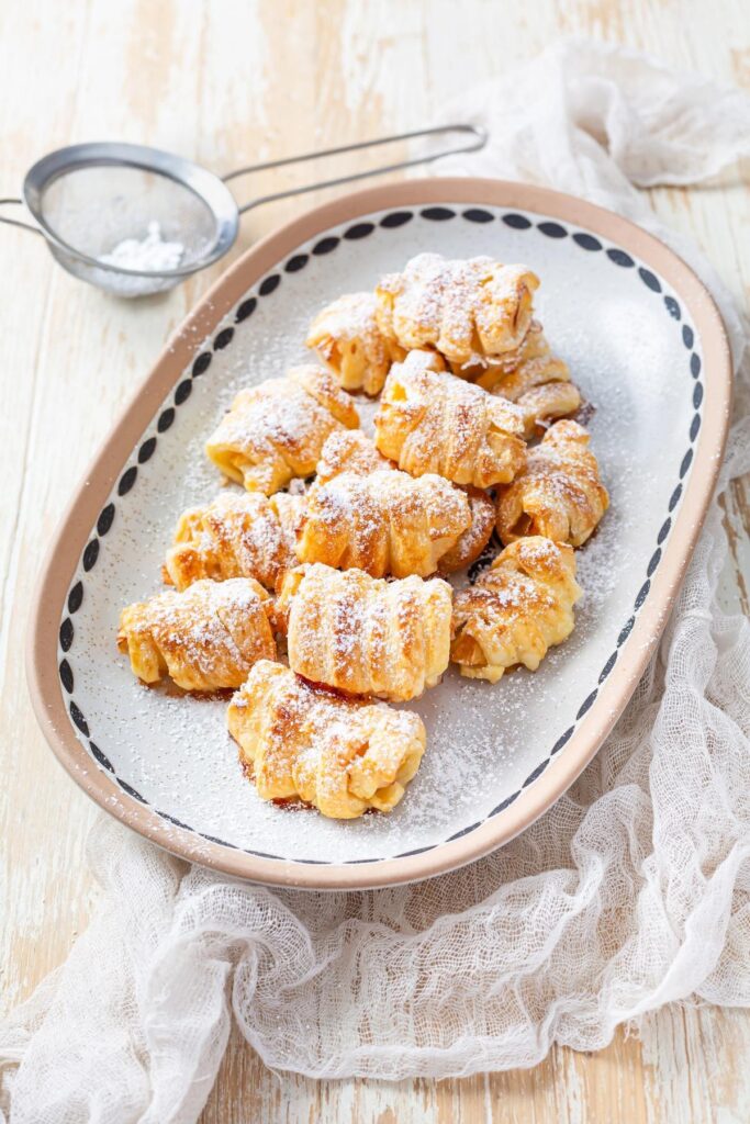 Flaky mini pastries are arranged on a decorative platter, dusted with powdered sugar for a cozy, homemade feel. These sweet bites are a charming and low-effort addition to your gathering spread.